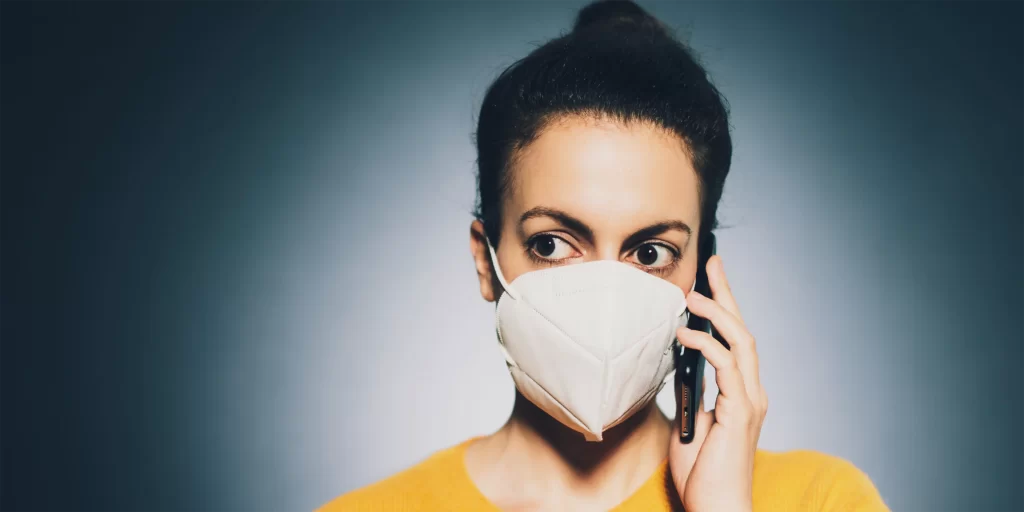 A person with olive skin and curly dark hair wears an N95 mask and a yellow sweater, holding a phone to their ear.