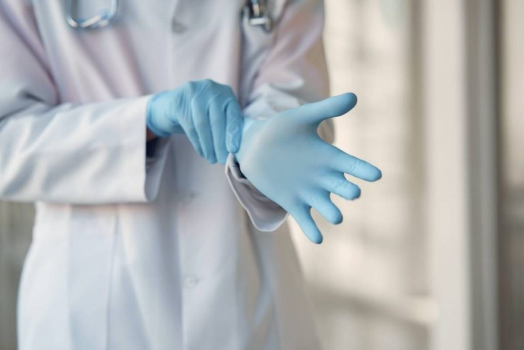 Closeup of a healthcare worker in a white lab coat donning a pair of blue medical gloves