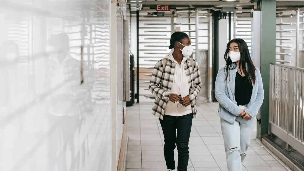 A Black person in a plaid shirt and an Asian person with long hair and a jean jacket, both wearing N95s, walk down a white corridor looking at each other.